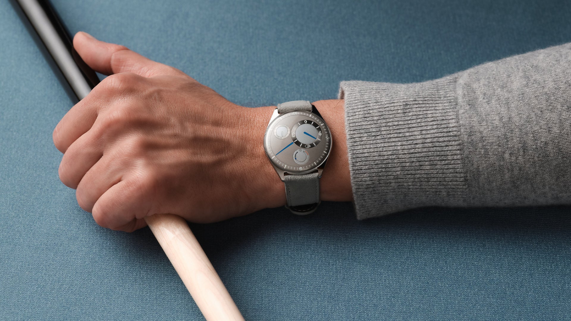 Hand wearing a Ressence TYPE 11 Latte, a contemporary luxury watch on a blue surface with a pool cue