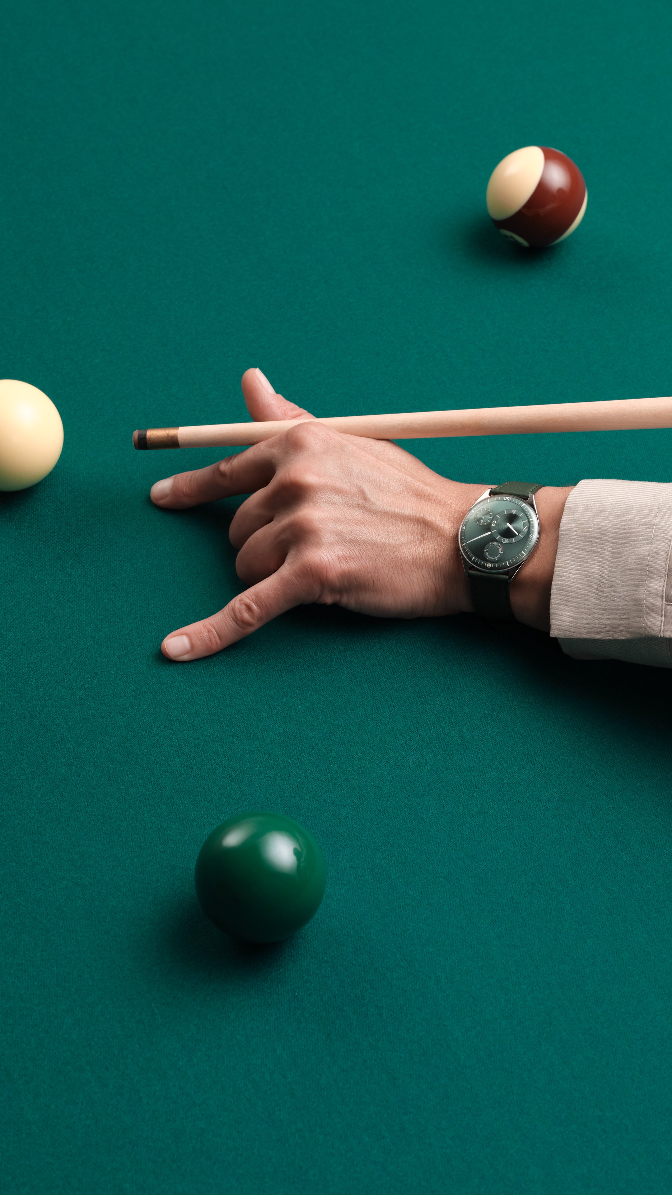 Vertical image of hand holding a pool cue approaching a ball on a green pool table wearing a Ressence TYPE 11 Pine, a contemporary luxury watch on the wrist