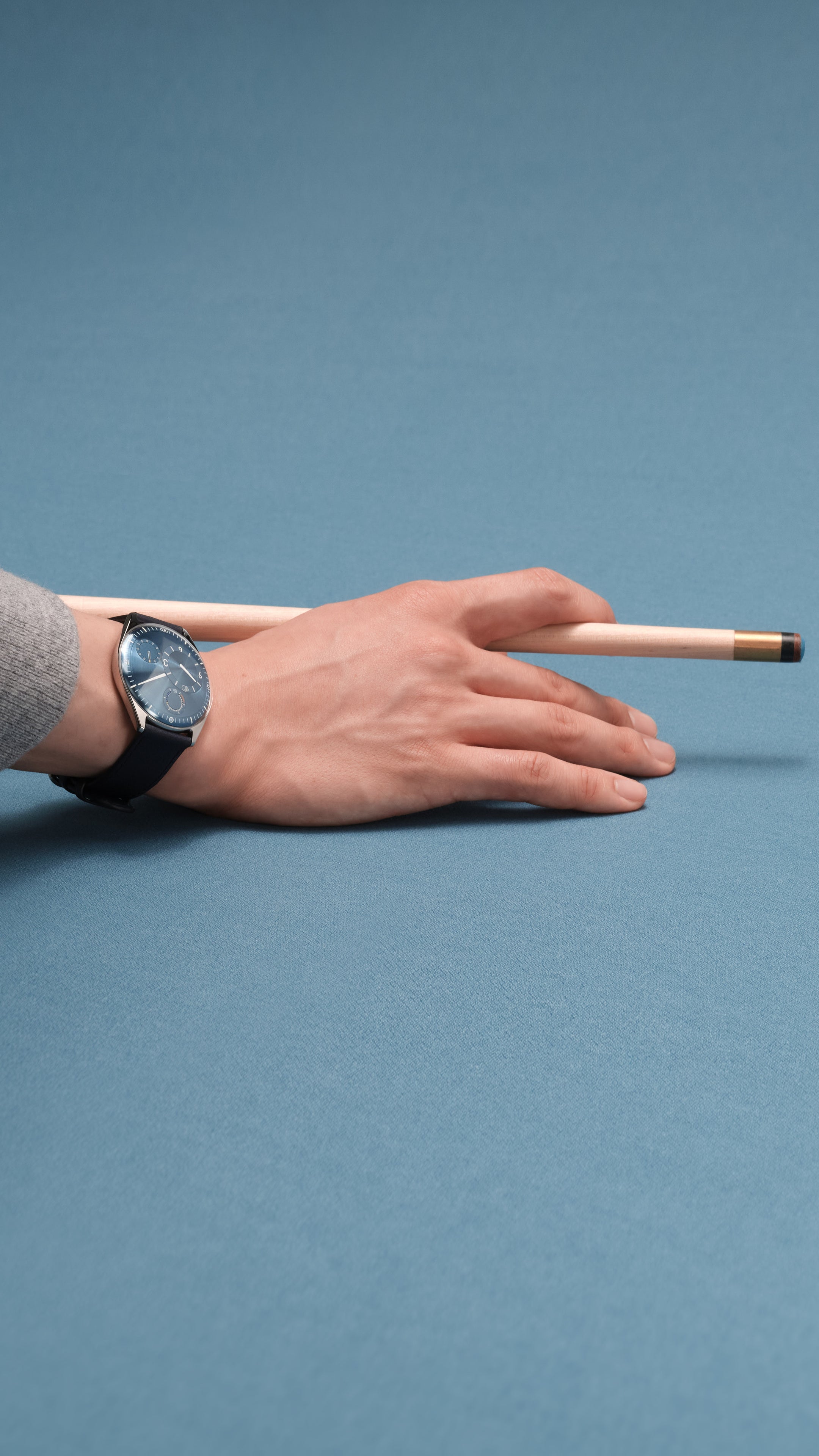 Vertical image of a hand holding a pool cue near a pool ball on a blue background wearing a Ressence TYPE 11 Sky, a contemporary luxury watch with a blue dial on the wrist