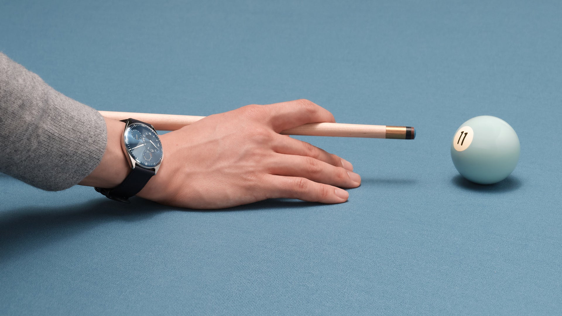 Hand holding a pool cue near a pool ball on a blue background wearing a Ressence TYPE 11 Sky, a contemporary luxury watch with a blue dial on the wrist