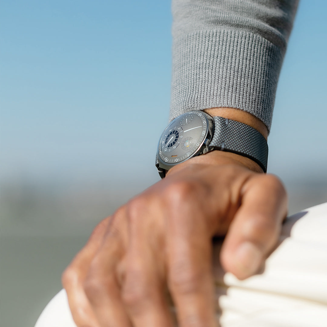 Vertical image of a person wearing a Ressence TYPE 2 connected watch with their hand resting on their knee with a blurred background