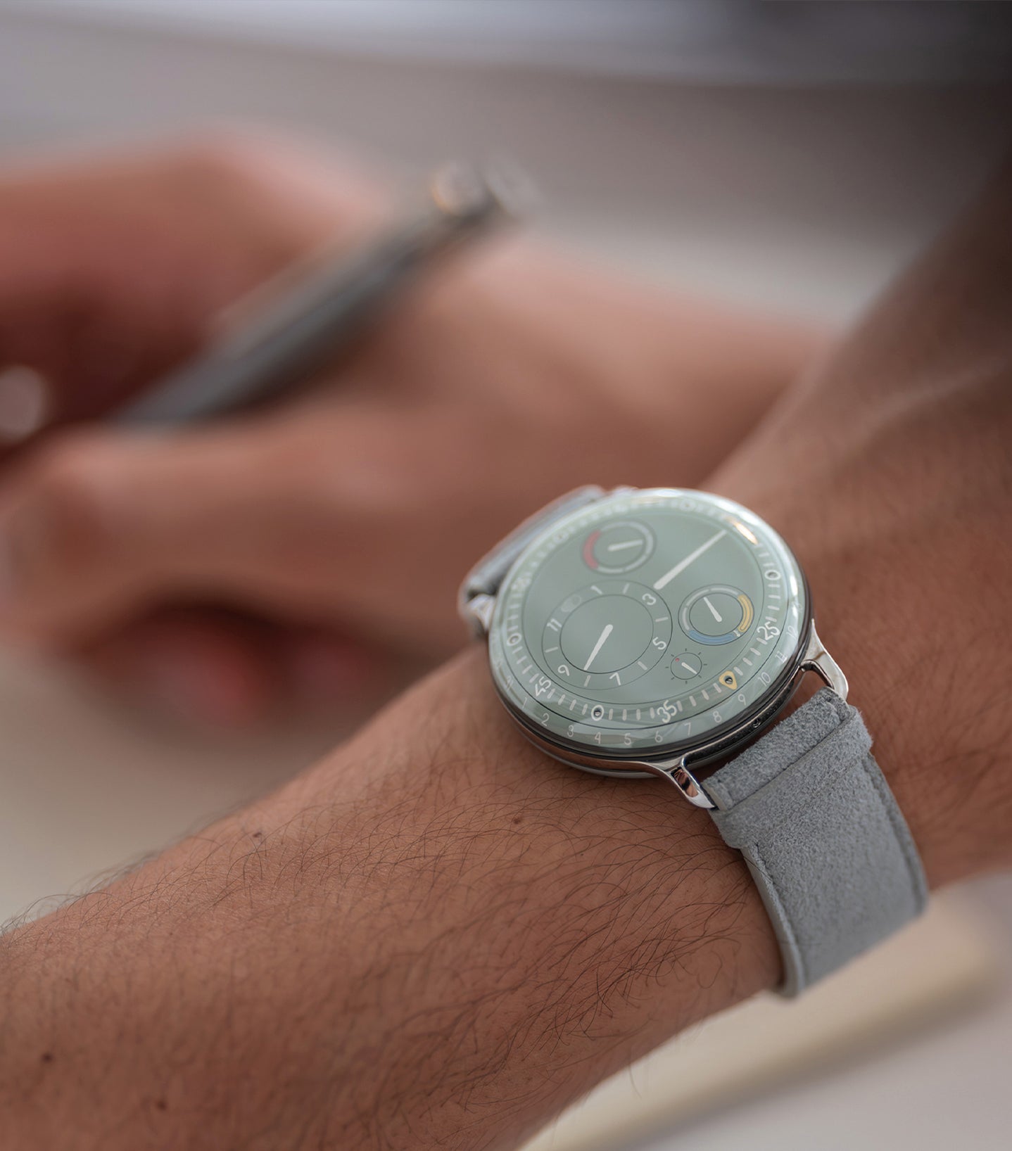 Ressence TYPE 3 Eucalyptus, the award-winning luxury oil-filled mechanical watch, shown on a man's wrist and holding a grey pen. vertical image.