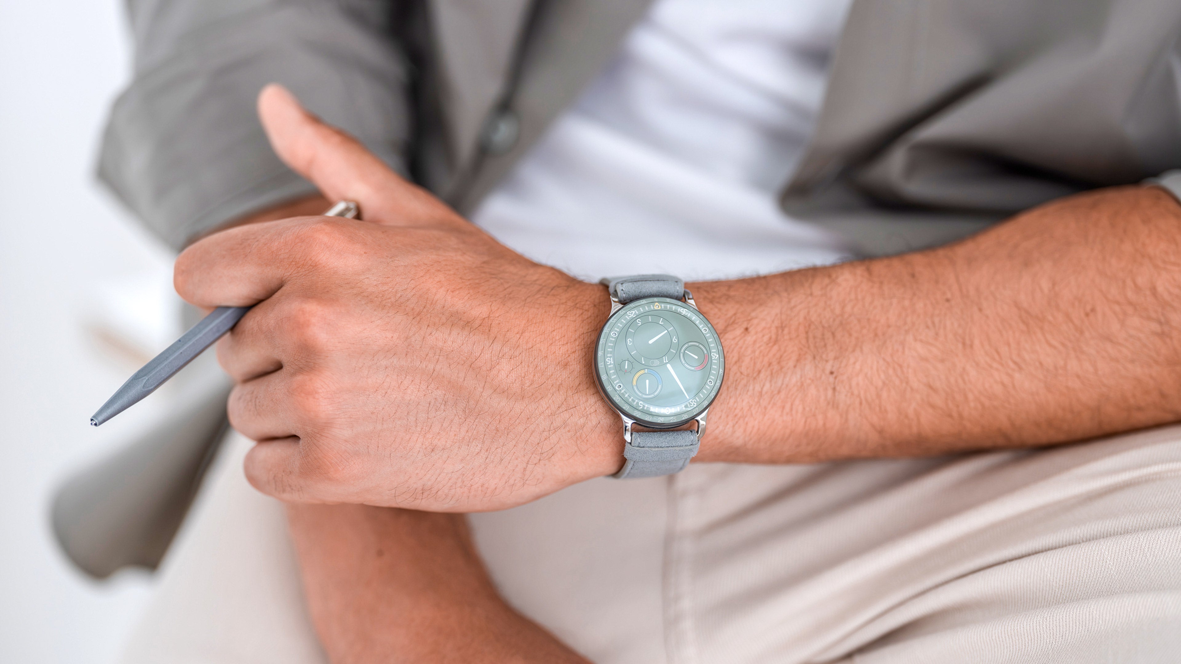 Ressence TYPE 3 Eucalyptus, the award-winning luxury oil-filled mechanical watch, shown on a man's wrist and holding a grey pen.