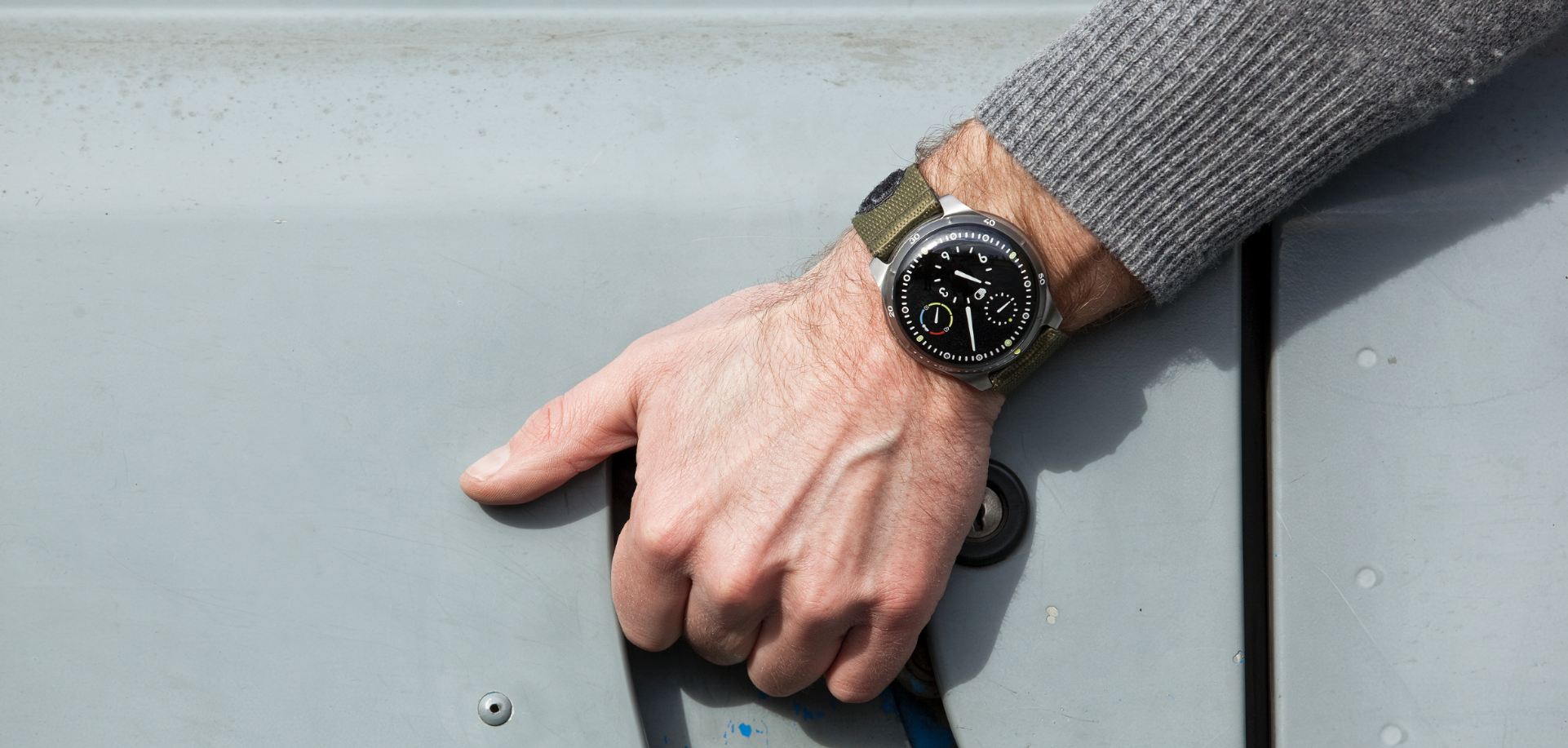 A man's hand grabbing a grey car door and wearing a Ressence TYPE 5 Black oil-filled diver's watch with black dial and grey nato strap.
