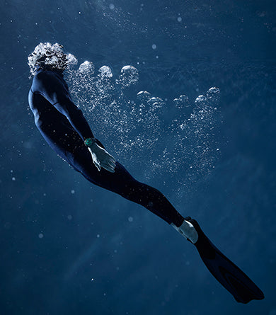 Vertical image of a diver going back to the surface, surrounded by air bubbles and wearing the Ressence TYPE 5 Luminova, an oil-filled diver watch with its dial fully filled with Super Luminova
