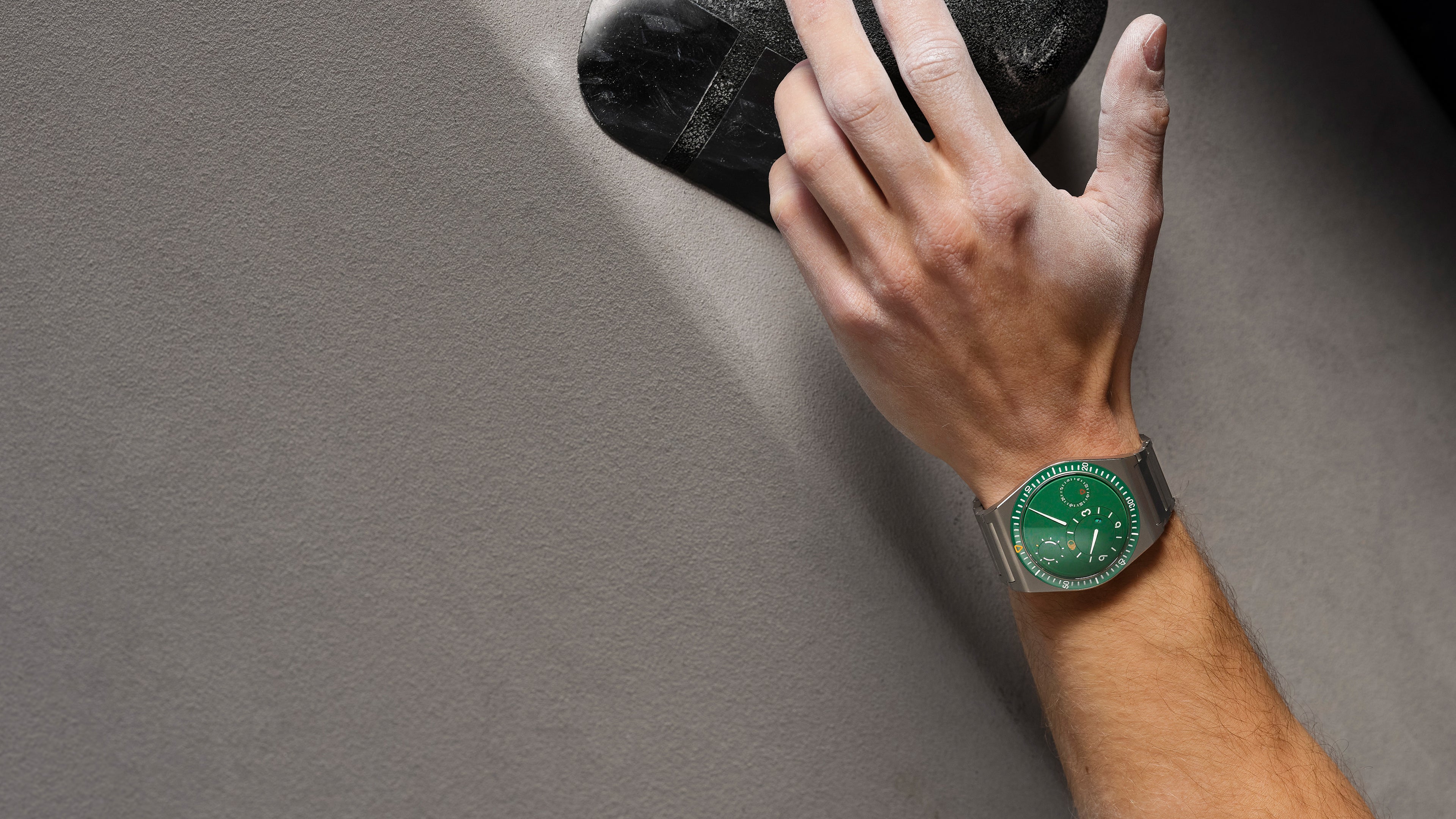 Hand wearing a cactus green Ressence TYPE 7 sport-chic GMT watch on a gray background