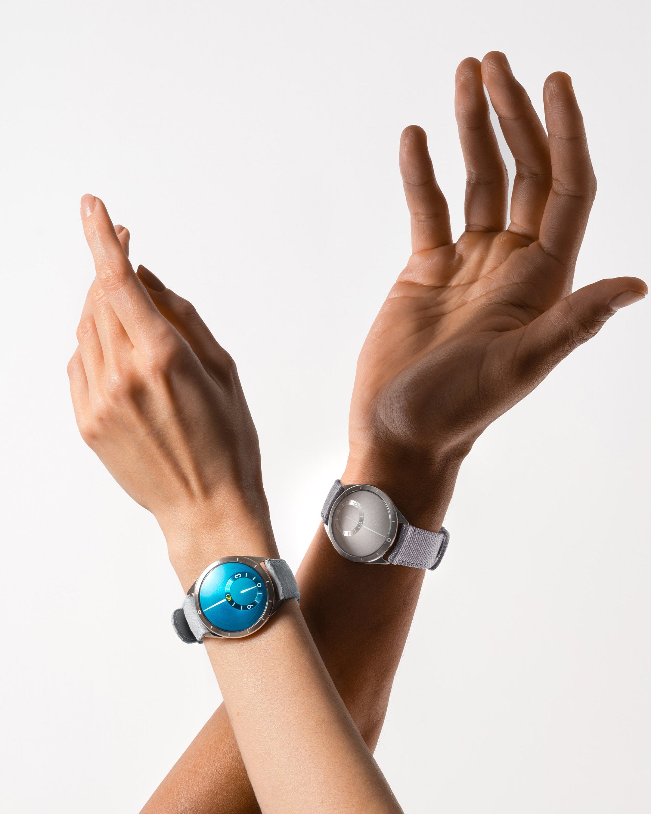 Two arms, one of a woman with a Ressence TYPE 9 Aqua and man's one with a Ressence TYPE 9 Grey both raised up, vertical image on a grey background